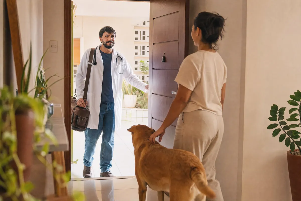 Rafael Holanda chegando à porta de um apartamento com a bolsa de atendimento, recebido pela tutora e pelo cachorro